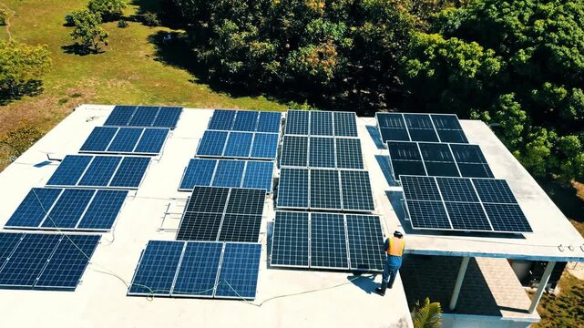 Aerial view of a worker in a yellow vest inspecting solar panels on a white rooftop surrounded by lush green trees in Guanaja, Bay Islands Department, Honduras.