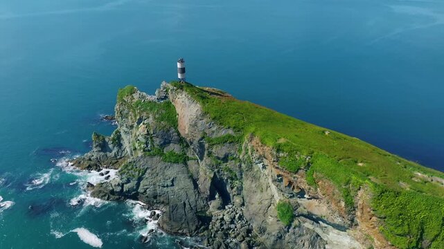 Drone footage of a striped lighthouse on a rugged green cliff above the calm sea in Vilyuchinskaya Bay near Vilyuchinskaya Sopka, Kamchatka Krai, Russia