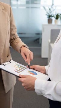 Two professional Caucasian business women in suits discussing financial charts and graphs on a clipboard in a modern office. Close-up of hands pointing to colorful pie and bar diagrams.