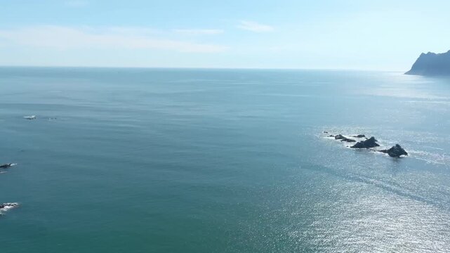 Drone footage of a striped lighthouse on a rocky green headland above calm blue sea, with mountain ranges on the horizon in Vilyuchinskaya Bay, Kamchatka Krai, Russia