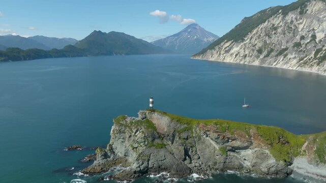 Aerial footage of a striped lighthouse on a cliff above calm sea in Vilyuchinskaya Bay, with Vilyuchinskaya Sopka volcano and mountains, Kamchatka, Russia