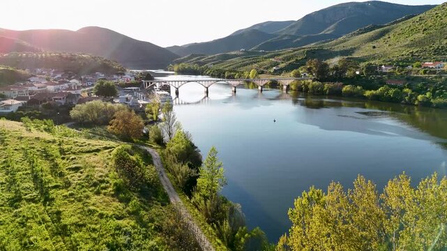Aerial view of Ponte Almirante Sarmento Rodrigues spanning the Douro River with lush green hills and a small village under bright sunlight Distrito da Guarda, Portugal.