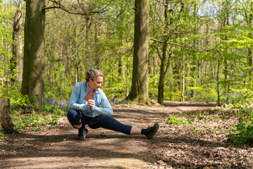 Obraz premium Woman stretches her leg while sitting on a path in a green forest during daytime in spring