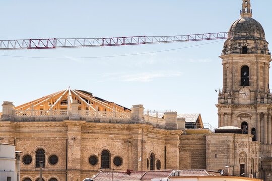Work on the roof of Malaga Cathedral, installing new tiles on a wooden structure, with a crane passing in front of its iconic single tower