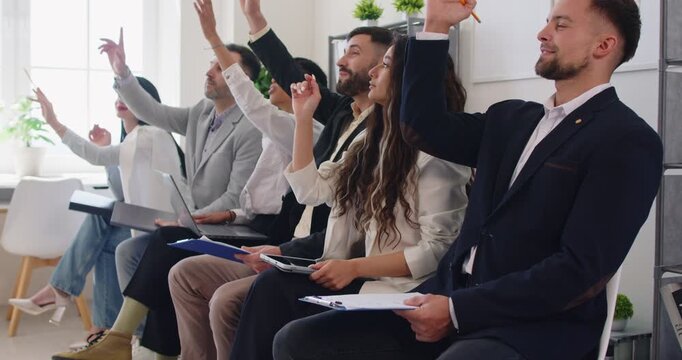 Diverse business team sitting in row and raising hands to ask or answer questions during work meeting. Engaged employees listening to speaker and taking notes while participating in discussion.