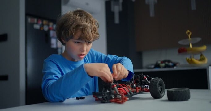 Young boy in a blue shirt carefully assembling and repairing a remote control car chassis with exposed wires and wheels on a table indoors