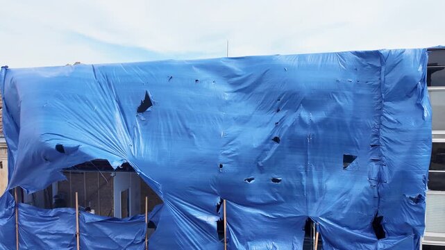 Tattered blue tarp billows in the wind over a building under construction. A large, torn blue tarp is stretched over a building undergoing renovation, flapping dramatically in the wind against a