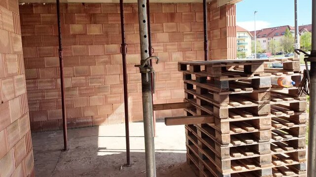 Construction site interior with scaffolding and brick walls. View inside a building under construction, showing brick walls, support poles, and stacked wooden pallets.