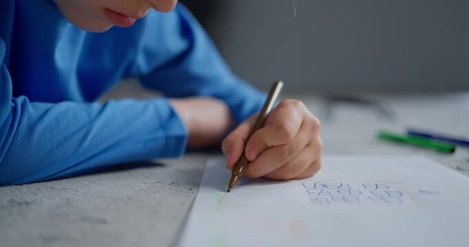 Young child in blue shirt lying at a table and concentrating while drawing with a pencil on paper in a quiet indoor learning moment