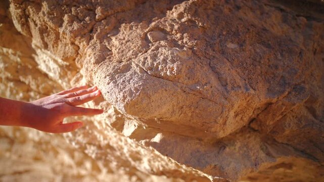 Close-up of a hand gently touching and feeling the textured surface of a rugged sandstone boulder bathed in golden sunlight. Warm orange and tan tones highlight the weathered rock formation.