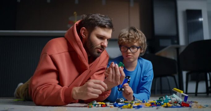 Father and young son lying on living room floor building colorful toy brick creations together, sharing focused playful learning time indoors