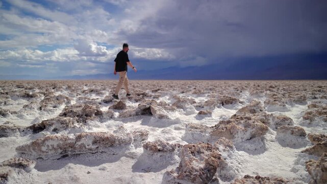 Man in black shirt walks across salt flats at Devils Golf Course with crystallized mineral formations under dramatic cloudy sky. Mountains visible on distant horizon, Death Valley National Park.