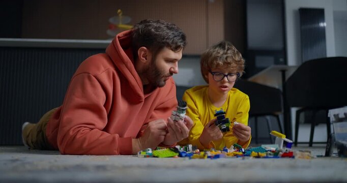 Father and young son lying on living room floor building colorful toy brick creations together, sharing focused indoor playtime and bonding