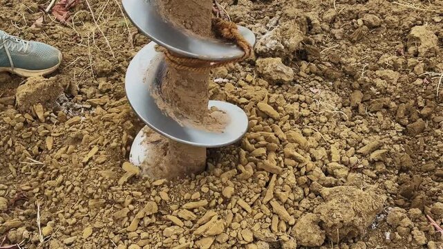 Close-up view of auger drill bit rotating into soil with loose earth around and worker using hand tool nearby, showing active drilling process in rural setting