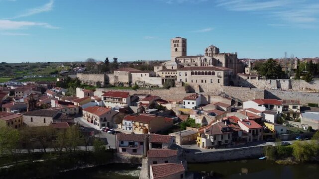 A majestic 4K drone video captures a full orbital flight around the medieval walled city of Zamora, Spain, on a sunny day. The camera sweeps over the robust stone fortifications, focusing on the Roman