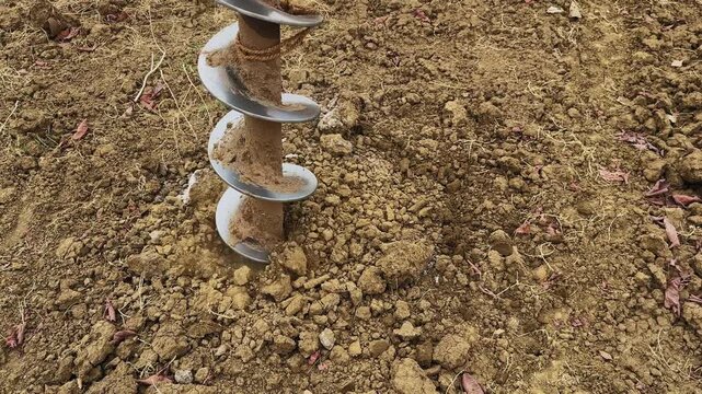 Mid view of auger drill bit partially inside ground with loose soil and gravel around, showing ongoing drilling process in a rural agricultural environment