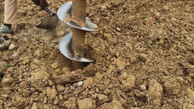 Close view of auger drill bit digging into soil while worker clears loose earth with hand tool, active ground drilling process in a rural agricultural setting