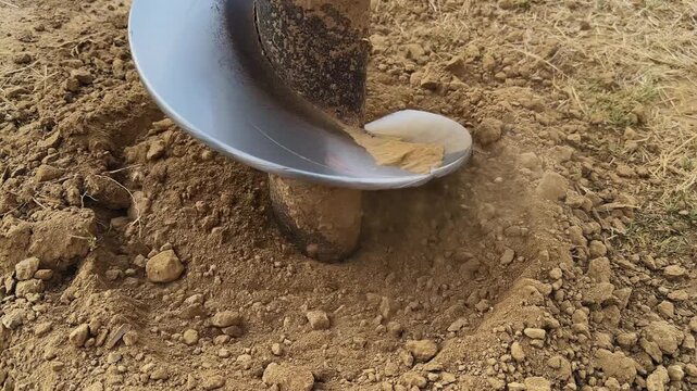 Extreme close-up view of auger drill bit penetrating ground with fine soil particles and small rocks displaced around the spiral blade during drilling process