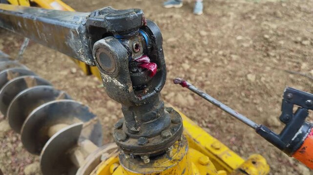 Close view of worker applying grease to universal joint of auger drive shaft, lubrication process on tractor attachment in a rural agricultural setting