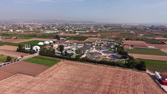 Modern wastewater treatment plant in Spain with circular clarifiers, biological nutrient removal, anaerobic digesters for biogas production, surrounded by agricultural fields near Mediterranean coast