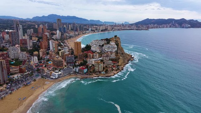 Benidorm Spain coastal cityscape with turquoise Mediterranean sea, sandy beaches, rocky headland, high-rise buildings and mountain backdrop under cloudy sky