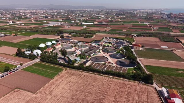 Modern wastewater treatment plant with circular clarifiers, biological treatment tanks, biogas digesters and green surroundings, viewed from above near coastline and farmland