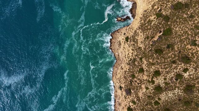 Top-down view of turquoise ocean waves crashing against a rugged rocky coastline with arid scrubland vegetation along the cliff edge