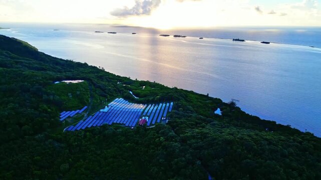Aerial view of a large solar panel array nestled in the lush tropical forest on a hillside overlooking the Caribbean Sea at sunset in Sandy Bay, Bay Islands Department, Honduras.