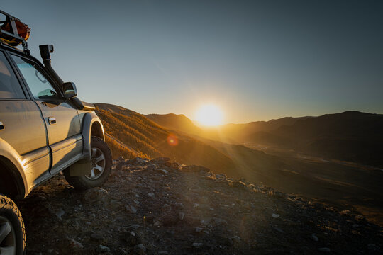 Rugged off-road 4x4 expedition vehicle parked on a rocky mountain edge during a beautiful golden hour sunset, overlooking a vast valley and distant peaks in a wild nature landscape scenery
