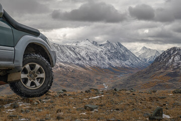 Close-up of a rugged 4x4 expedition vehicle with snorkel and mud terrain tire parked on a high mountain pass overlooking a vast snowy valley and jagged peaks under a dramatic overcast sky © ANDREY