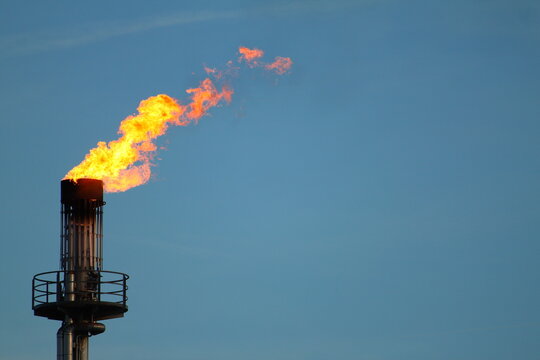 Industrial gas flare stack burning with bright orange flame against blue sky at petroleum refinery or chemical plant illustrating energy production and fossil fuel emissions. Copy space.