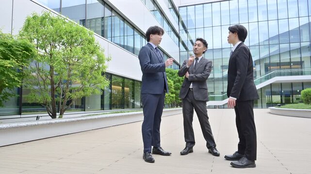 An image of three businesspeople in suits conversing outdoors in front of a modern office building; a full-body video of the corporate sales team communicating