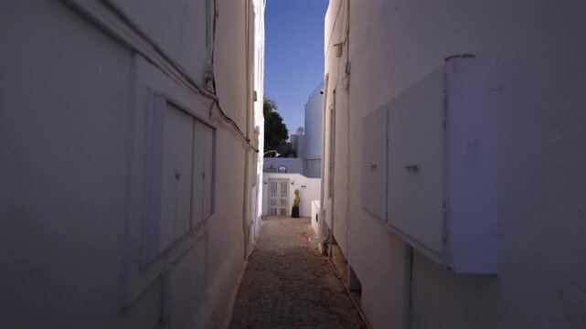 Camera moves through narrow alleyway between whitewashed walls toward white gate and courtyard. Cobblestone path leads past weathered doors under clear blue sky. Santorini, Greece