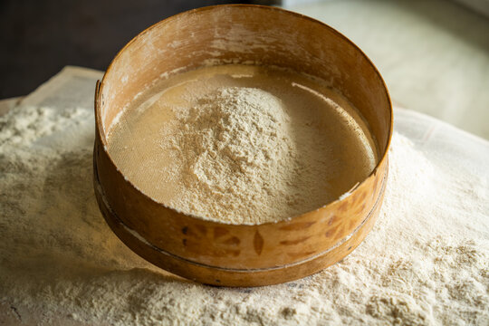 Vintage round wooden sieve containing mound of white wheat flour sits atop messy workspace covered in fine grain powder showcasing traditional home baking preparation and rustic culinary art
