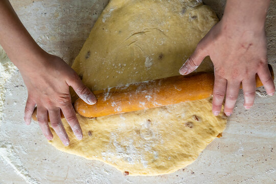Person presses timber tool against soft elastic yeast pastry containing raisins to flatten mass on flour covered marble countertop during traditional home baking process