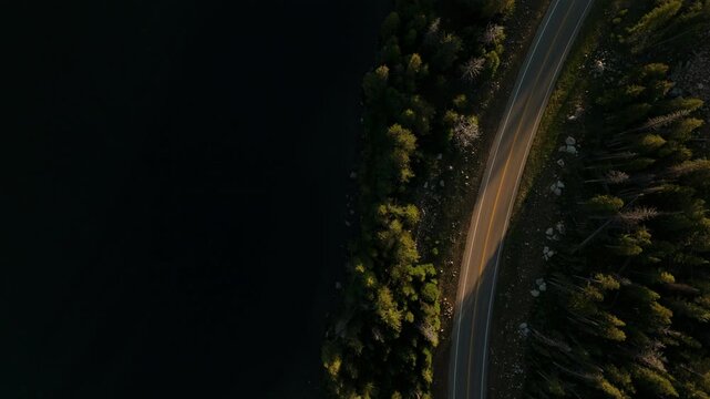 Drone footage captures a white car traveling along the scenic Beartooth Highway in Montana during sunset. The winding road cuts through dense evergreen forest beside a dark mountain lake.