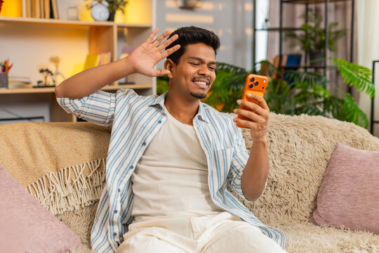 Indian man on home sofa waves at smartphone camera during video call greeting with friendly smile and chat. Arabian guy on home sofa speaks on screen then waves goodbye with warm casual mood indoors.