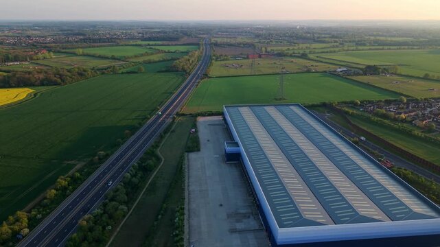 Aerial view of a large industrial warehouse next to a motorway surrounded by green fields and rural landscape during sunset in Doncaster, England, United Kingdom.