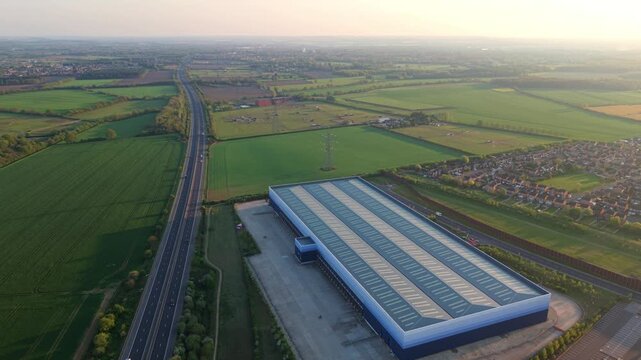 Aerial view of a large distribution warehouse and motorway surrounded by green fields and residential houses during sunset in Doncaster, England, United Kingdom.