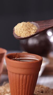 Close-up of brown cane sugar on a wooden spoon held over a traditional clay cup of tea. Authentic rustic setting highlighting natural sweeteners and cultural beverage preparation
