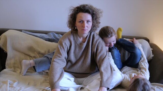 Tired mother tries to meditate on the sofa while her two energetic children jump and play wildly around her, capturing chaotic parenting and the struggle to find calm at home