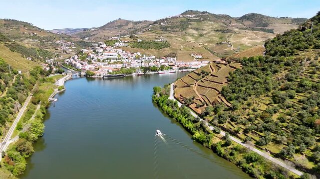 Aerial view of the Douro River with a boat cruising past the terraced vineyards and the historic town of Pinhao, Vila Real, Portugal.