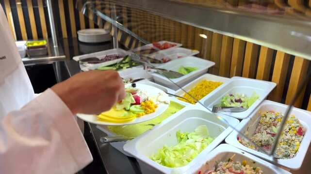 Guest in a white bathrobe selecting fresh vegetables and salads from a self service buffet line, carefully placing lettuce onto a plate full of healthy food options during breakfast at a hotel