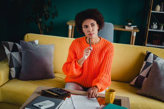 Young woman on yellow sofa thinking and budgeting at home with calculator, documents and coffee in cozy orange sweater