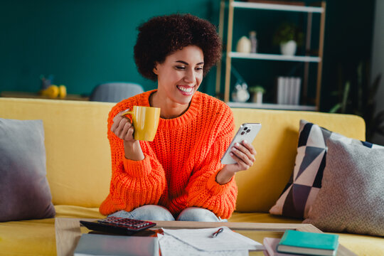 Smiling woman on a yellow sofa at home enjoying coffee while checking her smartphone, a relaxed casual moment in a colorful living room with notes and a calculator