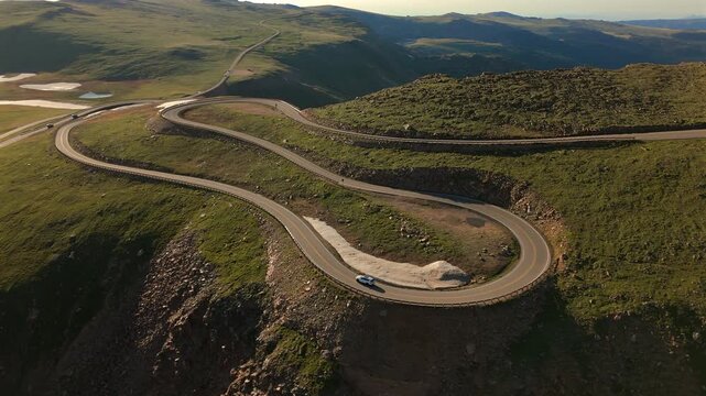 Drone footage captures Beartooth Highway in Montana as a vehicle navigates serpentine curves through alpine tundra with snow patches and distant lakes visible at golden hour.