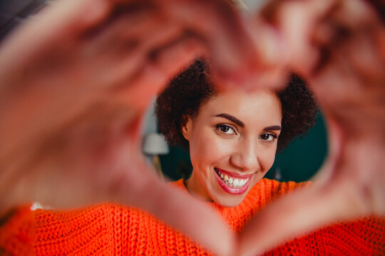 Smiling young woman making a heart with her hands while wearing a bright orange sweater and looking through the heart-shaped frame