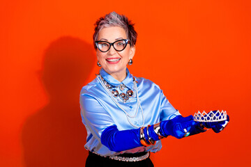 Confident mature woman in blue satin blouse and tiara smiles as she presents a sparkling crown against a bold orange backdrop © deagreez