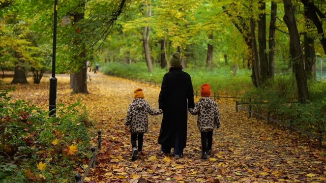 Rear view of a woman holding hands with her two young twin girls, walking together on a path covered with fallen yellow leaves during a peaceful stroll in an autumn park
