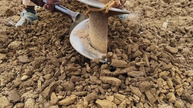 Close-up view of auger drill bit penetrating soil with loose earth and rocks surrounding, worker assisting with hand tool in an active drilling process outdoors